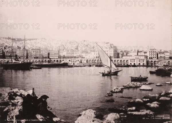 Alger, vue générale à l'entrée du port, View of the port of Algiers, photomechanical print, number: '1', address: '26 rue Bab-Azoun ALGER', photographer: Alexandre Leroux, Algiers, cliché maker: anonymous, c. 1875 - c. 1900, Alexandre Leroux, photographer, cliché maker, North Africa, pasteboard, paper, collotype, photoprint, height 247 mm x width 318 mm, harbour, A wide waterfront scene with calm water in the foreground and a rocky shoreline at the bottom edges, a small sailboat with a large triangular sail and a few people aboard positioned near the center right, a smaller rowboat with a seated person closer to the right edge, several larger moored ships and boats along the left and middle distances, a long quay with arched openings and multi-story buildings behind it stretching across the middle of the image, a densely built hillside of light-colored buildings rising behind the quay, and a person sitting on the rocks in shadow in the lower left foreground.