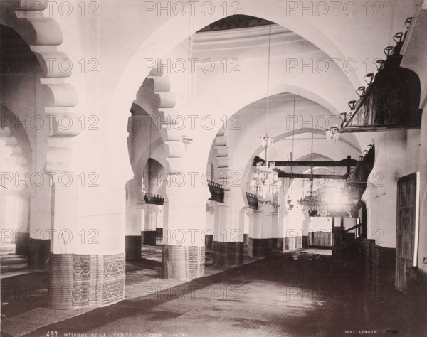 Interieur de la Mosquee El-Kebir-Alger, Interior of the Djama'a al-Kebir in Algiers, photomechanical print, number: '487', photographer: Alexandre Leroux, Algiers, cliché maker: anonymous, c. 1875 - c. 1900, Alexandre Leroux, photographer, cliché maker, North Africa, pasteboard, paper, collotype, photoprint, height 247 mm x width 318 mm, temple, shrine, Islam, Mohammedanism, Djama'a al-Kebir, A large interior space with a series of tall, rounded arches receding into the distance, each arch supported by thick columns whose lower portions are wrapped in patterned coverings, textured floor coverings laid across the ground, ornate hanging chandeliers suspended from the ceiling, pairs of decorative wooden or metal balcony-like structures attached to some arches, carved or scalloped arch details at the tops of several supports, a darker raised platform or structure along one side with mounted lights, high plain walls and vaulted ceilings, and a door opening visible at the far right.