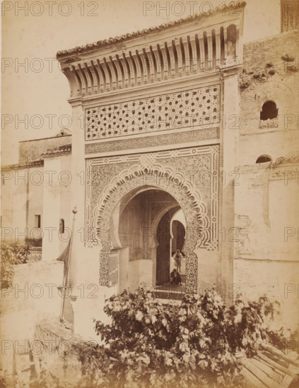 Mosquée Boumédine Tlemcen, Entrance of the Sidi Boumédienne mosque in Tlemcen, Algeria, photograph, inscription: 'Morocco', number: '234', Tlemcen, 1860 - 1900, photographer, North Africa, pasteboard, paper, albumen print, height 317 mm x width 247 mm, temple, shrine, Islam, Mohammedanism, Sidi Boumédienne mosque, A sepia-toned photograph of an ornate architectural façade featuring a large horseshoe-shaped arch with intricate geometric and floral tilework, framed by decorative panels and a carved band above. The upper section shows a lattice-like screen with repeating star patterns beneath a projecting cornice supported by a row of carved brackets. Flanking walls of plain plaster and brick rise on either side, one with small arched openings. Through the archway a passage and additional arches are visible, and two figures stand and sit near the entrance. In the foreground dense foliage and a low stone wall occupy the lower portion of the image.