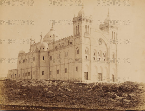 View of the Cathedral of Saint-Louis on the Byrsa in Tunisia, Cathédrale de Carthage, photograph, number: 'N. 122.', photographer: J. André Garrigues, Byrsa, 1884 - c. 1900, North Africa, pasteboard, paper, albumen print, height 247 mm x width 317 mm, church (exterior), Saint-Louis Cathedral, A large pale building with ornate towers and domes sits on a rocky, grassy foreground; the facade shows arched doorways and windows, decorative balustrades and carved details, narrow vertical openings in the towers, a circular rose window set within a recessed arch, and small pinnacles along the roofline.