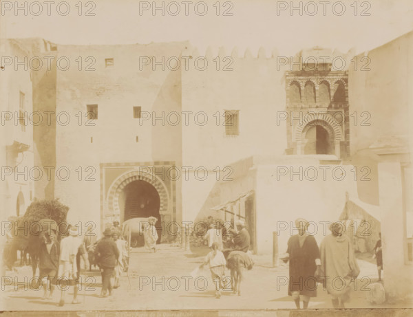 Cityscape in Sousse, Sousse, Porte de la Quarantaine, photograph, number: '276', photographer: Albert Goupil, 1860 - 1900, North Africa, pasteboard, paper, albumen print, albumendruk, height 247 mm x width 317 mm, passage, A pale street scene framed by tall, light-colored walls with small square windows and an ornate arched doorway set into a darker patterned façade, showing several people in loose garments and head coverings walking or standing in the foreground, animals including a donkey or mule and a larger pack animal near the central arch, stacked bundles or hay and a small pile of stones beside a low wall, a second arched entrance partially visible with striped archway decoration, and simple wooden awnings and poles casting soft shadows on the ground.