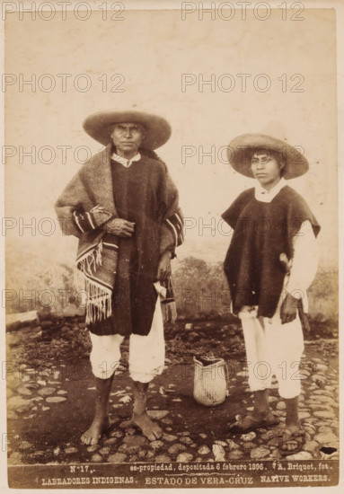 Labradores Indigenas, Native workers, Double portrait of two unknown inhabitants of Mexico, Estado de Vera-Cruz, photograph, copyright notice: 'es-propriedad, depositada 6 febrero 1896.', number: 'N. 17.', photographer: Abel Briquet, Veracruz, 1896, North America, pasteboard, paper, albumen print, height 194 mm x width 133 mm, height 210 mm x width 160 mm, adult man, An aged sepia photograph shows two males standing on a cobblestone ground in front of a plain wall; both wear wide-brimmed hats and loose garments layered over light-colored trousers, with the figure on the left draped in a fringed woven blanket and standing barefoot, the figure on the right wearing a dark poncho over a light shirt and sandals, and a woven basket placed on the ground between them.