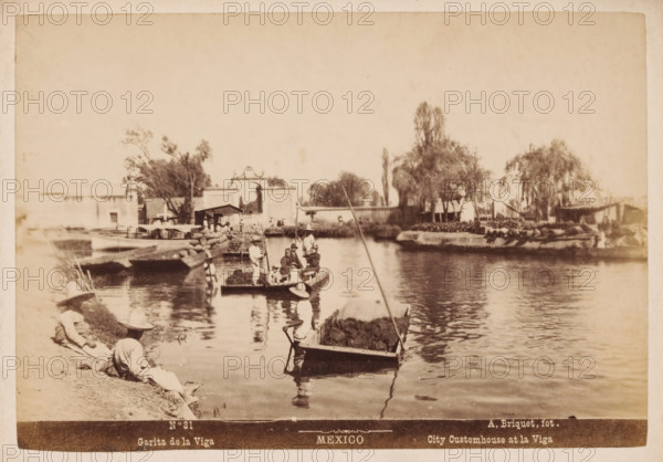Garita de la Viga, City Customhouse at la Viga, Pramen in the La Viga Canal in Mexico City, Mexico, photograph, number: 'N. 31', photographer: Abel Briquet, Mexico City, 1865 - 1910, North America, pasteboard, paper, albumen print, height 133 mm x width 194 mm, height 160 mm x width 210 mm, canals, waters (in city), city-gate, La Viga canal, A sepia-toned waterfront scene shows a calm body of water with several small flat boats, some loaded with sacks and vegetation, and people seated or standing in them wearing wide-brimmed hats and loose clothing; a few figures sit or recline on the grassy bank in similar attire. Along the opposite shore there are low buildings, a decorative arched gateway, trees with varying heights and foliage, and a narrow pier or dock where boats are moored. Reflections of the boats and people ripple on the water surface and a long pole extends from one vessel into the foreground. The overall image has a faded, aged appearance with soft contrasts and visible pape...