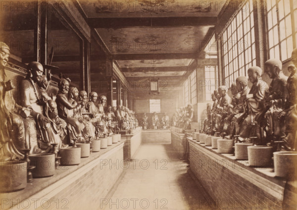 Interior of the Hualin Temple of Guangzhou with statues, photograph, annotation: 'Canton-Part of the Temple of the 500 Genii', 1850 - 1900, photographer, paper, pasteboard, albumen print, height 225 mm x width 310 mm, temple, shrine, traditional Chinese religions, Canton (city), A long interior corridor with rows of seated statues on raised platforms lining both sides, each statue placed on a round pedestal in front of a low brick wall; the statues depict robed human figures with varied headwear and hand positions, all facing inward toward the central walkway. The corridor has a high ceiling with decorative panels and hanging lanterns, tall multi-pane windows on the right side letting in light, square support columns and beams running along the length, and a distant window at the far end. The photograph is sepia-toned with visible wear, and the floor is a smooth tiled surface leading between the two lines of statues.