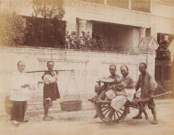 Five men with a wheelbarrow and carrying pole in Shanghai, photograph, annotation: 'Shanghai-Wheel barrow', Shanghai, 1850 - 1900, photographer, paper, pasteboard, albumen print, height 225 mm x width 310 mm, wheelbarrow, carrying something with carrying-pole, A sepia-toned street scene showing a low wall with decorative molding and a building façade behind it with columns, shutters, plants, and a gated entrance, in front of which five men stand and sit; one man on the left wears a light-colored tunic and dark trousers and stands next to a tall woven basket, another man holds a carrying pole with hanging baskets and wears a darker vest over lighter clothing, two men sit on a wooden cart with a large spoked wheel and one of them has a woven hat resting nearby, and a fourth man stands at the back of the cart holding its shafts, all wearing traditional loose garments and simple sandals, while foliage rises above the wall and architectural details such as pilasters and a pointed stone fini...