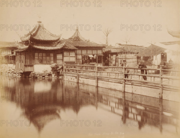 View of a temple the Shanghai, photograph, inscription: 'Sze Yuen Ming Co. 3 4, 42, Nanking Road', annotation: 'Shanghai. Chinese City', photographer: Sze Yuen Ming & Co, Shanghai, photographer: Sze Yuen Ming, 1850 - 1900, paper, pasteboard, albumen print, height 207 mm x width 258 mm, street (city(-scape) with figures, staffage), temple, shrine, traditional Chinese religions, A waterfront scene with ornate roofed buildings featuring curved eaves and tiled roofs, wooden framed windows, and lattice panels, a wooden railing running along a stone-lined water channel, reflections of the roofs and railing visible in the water, a few leafless trees rising above the rooftops, a sloped canopy supported by poles over a small area beside the railing, several people standing or leaning near the railing and under the canopy, simple wooden structures and fences behind the railing, stone and wooden elements at the water's edge, and muted sepia tones throughout the image.