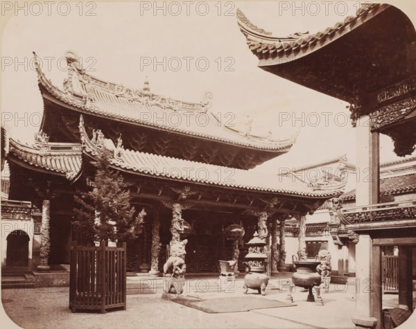 View of what is likely the Chenghuang Temple of Shanghai, photograph, annotation: 'Shanghai-City Temple', Shanghai, 1850 - 1900, photographer, paper, pasteboard, albumen print, height 230 mm x width 301 mm, temple, shrine, traditional Chinese religions, A courtyard scene of an ornate traditional building with sweeping tiled roofs and upturned eaves decorated with carved figures and patterns, carved columns topped by dragon sculptures supporting the roof, stone lion statues on pedestals near the entrance, large metal incense burners and vessels placed on the courtyard floor, a wooden slatted enclosure in the foreground, carved beams and decorative brackets under the eaves, a tree partially obscuring part of the structure, and additional architectural details and rooftops visible at the edges of the frame.