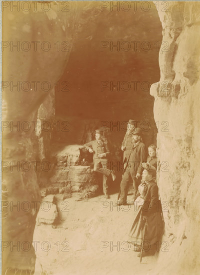 Tourists at the entrance of the Kohlscheuer cave near Echternach, Luxembourg, photograph, inscription: 'C.23, Environs d'Echternach., Kohlscheuer, grotte près d'Echternach', photographer: Henri van den Broeck, Echternach, 1890 - 1910, pasteboard, paper, daylight gelatin silver print, height 268 mm x width 209 mm, tourism, exploring caves, A sepia-toned photograph showing a group of people standing and sitting on a rocky ledge inside a cave or rock overhang, with rough textured rock walls visible on the right and left. Four adults in period clothing are visible, including men wearing suits and hats and women in long dresses, one woman standing with a walking stick and another seated on the ledge. One man leans against the rock with a hand resting on a small box beside him, and another man stands nearby with a cane. The ground is uneven and sandy, and the scene is framed by large rock formations that create a shaded interior area behind the group.