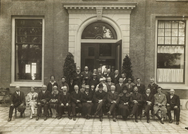 Group portrait of men and women in front of the entrance of a building, photograph, stamp: 'Copyright. Foto pers Bureau Hollandia Alkmaar, telephone 760', photographer: Hollandia, Foto Pers Bureau, Alkmaar, 1910 - 1930, paper, gelatin silver print, height 170 mm x width 235 mm, height 296 mm x width 355 mm, anonymous historical persons portrayed in a group, in a group-portrait, A large group of people arranged in rows in front of a brick building with an arched doorway and large rectangular windows, with two tall potted plants flanking the entrance; the men wear suits with ties or bow ties and many wear hats, some hold canes, and the women wear dresses or coats with cloche or brimmed hats, seated and standing on a paved courtyard with a bicycle visible at the left edge.