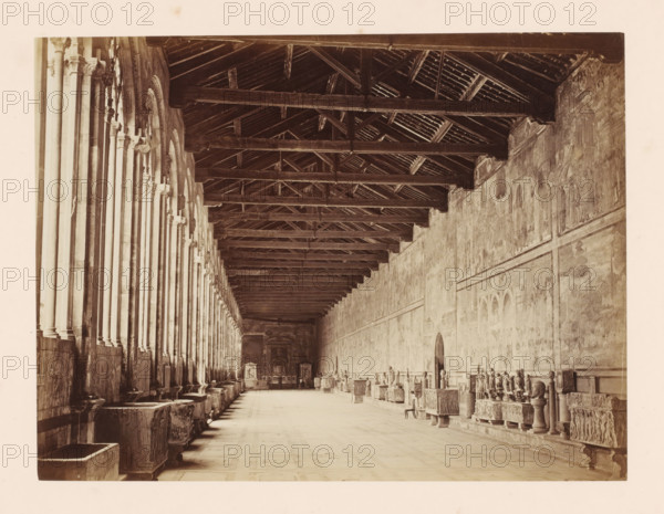 Gallery in the Camposanto in Pisa, Italy, Campo Santo. Galleria delle Cappelle, Pisa, photograph, address: 'GIACOMO BROGI, FOTOGRAFO-EDITORE, FIRENZE, LUNG ARNO DELLE GRAZIE 15', number: '3373', photographer: Giacomo Brogi, 1864 - 1881, Tuscany, pasteboard, paper, albumen print, height 347 mm x width 473 mm, gallery (spacious corridor), Campo Santo (Pisa), A long, vaulted interior corridor with a wooden roof structure of exposed beams and rafters running the length of the space, a row of tall stone columns with arched openings along the left side, a continuous stone wall along the right side covered with faint wall paintings or frescoes, and a tiled floor stretching toward a distant doorway and altarpiece; stone benches, sculpted sarcophagi, and carved architectural fragments line both sides, and a single chair sits near the right side.