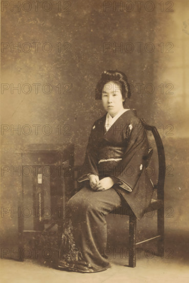 Portrait of an unknown Japanese woman, photograph, inscription: 'S. Hachiya. Kiriu. Gunma.', photographer: S Hachiya, Kiryu, c. 1890 - c. 1920, Gunma, paper, pasteboard, baryta paper, daylight gelatin silver print, height 137 mm x width 92 mm, height 228 mm x width 138 mm, adult woman, anonymous historical person depicted, A sepia-toned portrait of a seated woman wearing a traditional dark kimono with a lighter collar and patterned hem, her hands resting in her lap, hair styled in an updo, seated on a wooden chair next to a small wooden table, set against a mottled backdrop with visible age marks and surface wear.