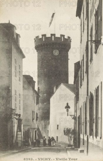View of the Tour de l'Horloge in Vichy, Vichy.-Vieille Tour, photomechanical print, stereograph, number: '8293.', Vichy, c. 1890 - c. 1910, photographer, pasteboard, paper, collotype, photoprint, height 138 mm x width 179 mm, tower, clock tower, monumental adornment, A narrow street flanked by tall buildings leads to a large round stone tower with battlements and a flagpole extending above it, the tower showing a circular clock face; the street scene includes a few people standing and walking near the base of the tower, a set of exterior stairs on the left side, shuttered windows on the building facades, an ornate wall-mounted street lamp on the right, and a small tree or shrub near the right sidewalk.