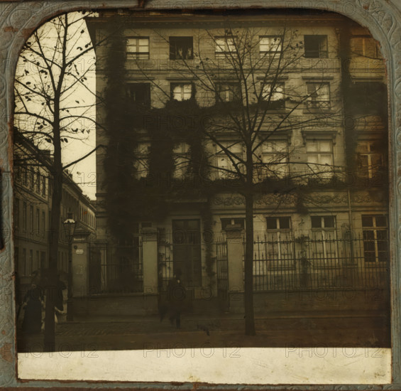 Part of the facade of the Richmodishaus in Cologne, Richmodis-Haus in Coln, stereograph, number: '575.', Cologne, 1900 - 1940, photographer, North Rhine-Westphalia, paper, plastic, pasteboard, organic material, inorganic material, cellulose nitrate film, metal, gelatin silver print, developing gelatin silver print, height 75 mm x width 150 mm, facade (of house or building), Richmodishaus, A faded sepia-toned photograph shows the facade of an older multi-story building with rectangular windows, some framed by climbing ivy and partially obscured by bare tree branches in the foreground; an iron fence with stone pillars runs along the front, and shadowy figures move along the sidewalk, their forms blurred; a narrow street and neighboring buildings flank the left side, and a streetlamp and a vertical signpost are visible near the corner.