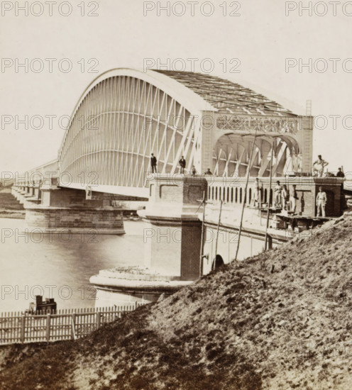 View of the Kuilenburg railway bridge near Culemborg, Bridge at Kuilenburg, stereograph, photographer: Pieter Oosterhuis, Culemborg, before 1868-09-06, pasteboard, paper, albumen print, albumendruk, height 86 mm x width 177 mm, bridge in village across river, canal, etc. - II - ideal landscapes, Kuilenburg railway bridge, A large arched metal bridge spans across water with a series of vertical and diagonal lattice supports forming the arch and deck, resting on stone piers rising from the river; workers stand and sit on the bridge structure and on the brick abutment, some near scaffolding and ladders, while wooden poles and construction materials are visible on the walkway; a grassy embankment slopes down toward a picket fence in the foreground, and calm water reflects parts of the bridge and piers.