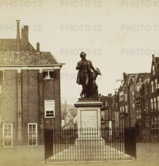 Ary Scheffer, Dordt, Statue of Ary Scheffer in Dordrecht, stereograph, Dordrecht, after sculpture by Cornelia Scheffer-Lamme, 1873 - 1890, Cornelia Scheffer-Lamme, photographer, after sculpture by, pasteboard, paper, albumen print, height 84 mm x width 170 mm, Person, monument, statue, A bronze statue of a robed man stands on a tall rectangular stone pedestal behind a metal fence, the man holding a hat in one hand and a small object near his chest with the other, flanked by stepped bases and finial-topped fence posts; surrounding buildings with brick facades and tiled roofs appear on both sides, with windows and chimneys visible, and a narrow street of multi-story row houses recedes into the background.