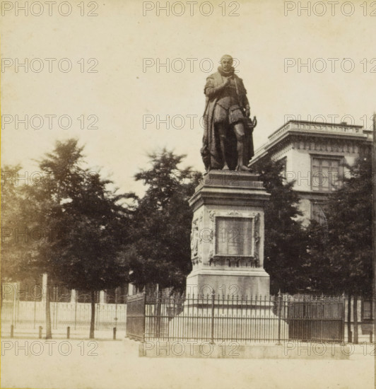 The Hague, Plein, View of the statue of William of Orange on the Plein in The Hague, stereograph, after sculpture by Louis Royer, 1873 - 1890, Louis Royer, photographer, after sculpture by, pasteboard, paper, albumen print, albumendruk, height 85 mm x width 169 mm, Person, Willem I (Prince of Orange), monument, statue, square, circus, A monochrome photograph showing a large statue of a robed figure standing on a tall, decorated stone pedestal behind an iron fence, with trees partially obscuring the lower part of the monument and a multi-story building with rectangular windows visible in the background, all set against a pale sky.