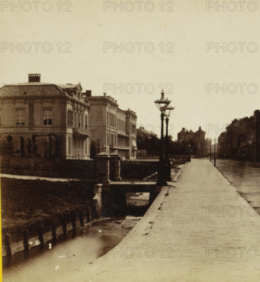 Zeestraat seen from Javastraat in The Hague, View of the Zeestraat seen from Javastraat in The Hague, stereograph, inscription: '(large building at the back left is a so-called 'rich people's courtyard')', The Hague, 1873 - 1890, photographer, pasteboard, paper, albumen print, height 84 mm x width 170 mm, street, canals, waters (in city), A sepia-toned street scene showing a wide sidewalk running into the distance alongside a narrow canal, with ornate multi-story buildings on the left featuring shuttered windows and decorative facades, a row of trees and a low fence along the canal, a stone bridge with balustrade crossing the water, tall vintage street lamps with multiple lanterns positioned near the bridge, and distant buildings and smaller lamps lining the far end of the street under an empty sky.