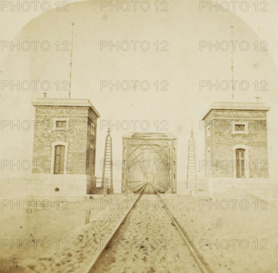 Netherlands, Great railway bridge at Moerdijk, completed 1871, View of the great railway bridge at Moerdijk, stereograph, Moerdijk, 1871 - 1872, photographer, pasteboard, paper, albumen print, height 86 mm x width 174 mm, railway, tramway, rack railway, bridge, Moerdijk railway bridge (1872), A sepia-toned scene showing a straight set of railroad tracks leading toward a metal truss bridge framed by two brick towers with rectangular windows and flat roofs, ladders and decorative finials flanking the bridge entrance, gravel and scattered debris along the track foreground, and a large pale sky occupying the upper portion of the image.