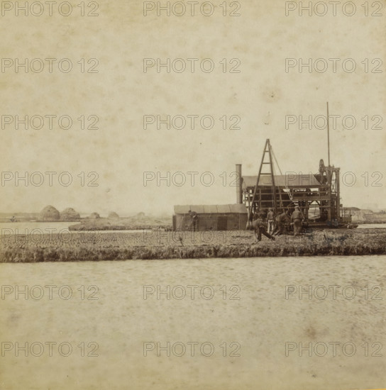 View of a state peat press machine in the low moor southeast of Amsterdam, In the low moor, Collection A, Geographical sculpture gallery, collected by MJ Broekhuijsen, headmaster of a school in Amsterdam, stereograph, inscription: SE of Amsterdam. The state peat press machine extracts the peat, presses it, and cuts peat blocks from it. See pl. 24.', number: 'A.23.', number: '23.', Amsterdam, publisher: Merkelbach & Co., compiler (person in literature): Martien Johan Broekhuijsen, 1900 - 1902, Merkelbach & Co., Martien Johan Broekhuijsen, photographer, publisher, compiler (person in literature), pasteboard, paper, albumen print, albumendruk, height 87 mm x width 176 mm, peat-cutting, world fair, world exhibition, A sepia-toned scene showing a body of water in the foreground and a narrow strip of land with grassy edges above it, behind which a working structure stands near the right edge; the structure has a tall vertical pole, a ladder-like frame, a small roofed section, visible machine...