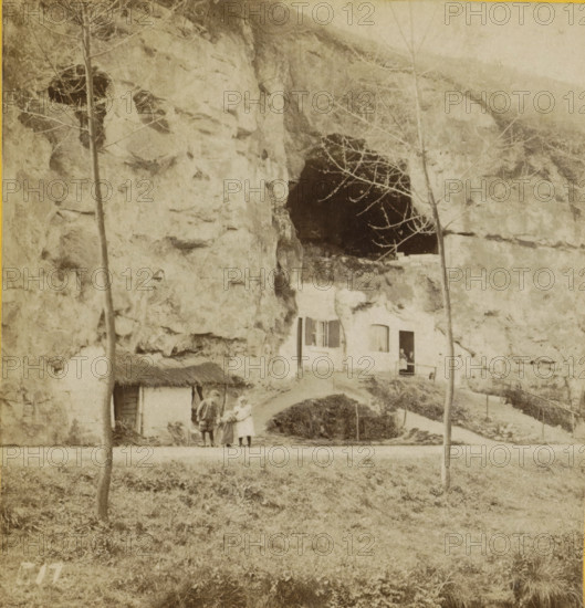 View of rock houses in Geulem, Limburg, Rock houses, Geographical sculpture gallery, stereograph, number: 'F.17.', inscription: '37', inscription: 'te Geulem, (see pl. 16.) excavated in the soft stone. Warm and dry, but ... poorly ventilated!', number: '17.', Geulhem, publisher: Merkelbach & Co., Amsterdam, compiler (person in literature): Martien Johan Broekhuijsen, 1873 - 1890, photographer, compiler (person in literature), pasteboard, paper, albumen print, albumendruk, height 87 mm x width 176 mm, cave-dwelling, A faded sepia photograph showing a rocky cliff face with a large dark cave opening near the top and a smaller dark recess to the left, two tall bare trees in the foreground rising in front of the rock, a small building built into the rock with a window and an open doorway and a woman standing in the doorway, a thatched-roof shed at the base of the cliff, three people standing on a path in front of the rock one wearing a dress and apron and another in a coat and hat, grassy g...