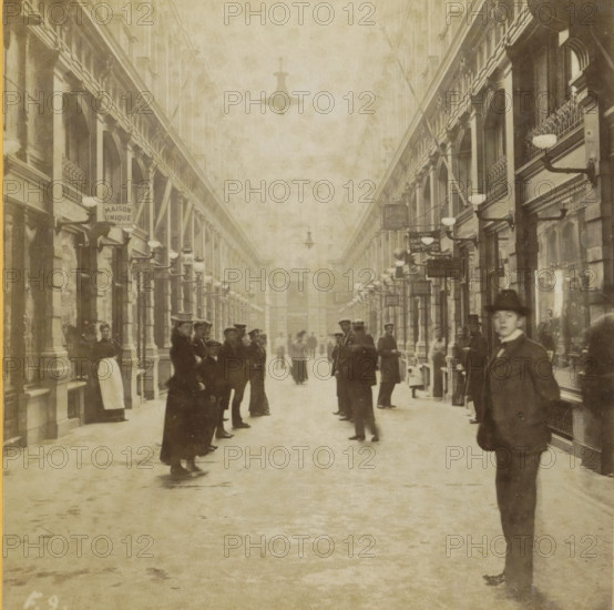 Covered shopping arcade de Passage in The Hague, Passage, Collection F, Geographical sculpture gallery, collected by MJ Broekhuijsen, headmaster of a school in Amsterdam, stereograph, inscription: 'in The Hague: covered shopping street. We see only half of it here, as the street curves at the end. Above us in the middle electric lighting.', number: '9.', number: 'F.9.', The Hague, publisher: Merkelbach & Co., Amsterdam, compiler (person in literature): Martien Johan Broekhuijsen, 1900 - 1902, photographer, compiler (person in literature), pasteboard, paper, albumen print, albumendruk, height 87 mm x width 176 mm, shop-window, show-window, arcade, architecture, A long covered passageway flanked by tall colonnaded facades with arched openings and decorative balconies lined with wall-mounted lamps, a high ceiling with hanging light fixtures, and numerous people standing and walking along the central floor; several individuals wear coats and hats, one person in the foreground on the right ...