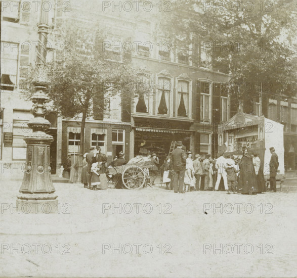 The Hague, Street scene with puppet show in The Hague, stereograph, inscription: 'RBK', inscription: '(puppet show ...)', photographer: attributed to Wilhelm Frederick Antonius Delboy, 1884 - 1928, Attributed to, paper, pasteboard, daylight collodion silver print, height 87 mm x width 180 mm, urban housing, street, puppet and marionette show, A sepia-toned street scene shows a row of multi-story buildings with tall windows framed by shutters and decorative moldings, partially obscured by leafy trees; an ornate lamp post with carved details stands in the foreground left; a market or gathering occupies the sidewalk with a wooden cart loaded and a group of adults and children clustered nearby, some wearing hats and long garments and a few women in high-collared dresses; a small painted booth or kiosk with decorative trim is visible at right; shopfronts with awnings and window displays line the ground level behind the crowd.