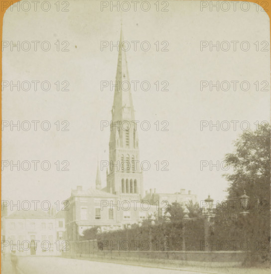 La Haye. Nouvelle Eglise Catholique, View of the Sint-Jacobus de Meerkerk in The Hague, Vues de Hollande, stereograph, number: '16.', The Hague, publisher: J. Lévy & Cie, Paris, 1878 - 1890, photographer, pasteboard, paper, albumen print, albumendruk, height 87 mm x width 178 mm, parts of church exterior and annexes, Parkstraat, Sint-Jacobus de Meerkerk, A tall church steeple rises above a cluster of buildings, the steeple showing narrow vertical openings and a pointed spire with visible structural details, set against a pale sky. In front of the church are multi-story rectangular buildings with rows of windows and simple façades. A row of trees with dense foliage lines one side, partially obscuring a decorative lamppost. A low stone wall and an iron fence run along the foreground beside a paved street. The image has a faded, sepia-toned appearance and rounded corners at the top.
