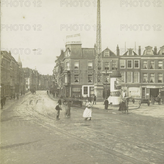 s Gravenhage.- Wagenplein, View of the Wagenplein in The Hague, stereograph, number: '112.', The Hague, 1882 - 1920, photographer, paper, pasteboard, gelatin silver print, developing gelatin silver print, height 89 mm x width 178 mm, urban housing, street, square, circus, Wagenstraat (The Hague), A wide street scene with tracks running along a paved roadway flanked by multi-story brick buildings, a tall lattice tower rising beside a corner building, an advertising column near the sidewalk, horse-drawn carts and a few figures walking or standing on the street, some people in period clothing with long skirts and coats, windows and dormers visible on the building facades, a distant row of buildings receding down the street, and a small airplane visible in the sky.
