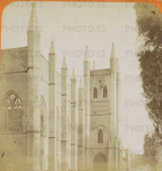 View of the Willemskerk in The Hague, La Haye. Église Guillaume, Vues de Hollande, stereograph, number: '18.', The Hague, publisher: Léon & Lévy, Paris, publisher: J. Lévy & Cie, 1873 - 1890, photographer, pasteboard, paper, albumen print, height 87 mm x width 178 mm, parts of church exterior and annexes, Willemskerk (The Hague), A faded sepia image of a large Gothic-style stone building with tall narrow buttresses topped by pointed pinnacles, arched windows with tracery, a tower section with louvered openings and a circular clock face, and a dark arched doorway at ground level, with a large dark tree partially visible along the right edge.