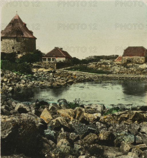 Bornholm. Christianshöh, Forts on Christiansø, near Bornholm, stereograph, number: 'S 24987', monogram: 'BW', inscription: 'Dänemark. Bornholm. Christianshöh, Denmark. Bornholm. Christianshöh., Dinamarca. Bornholm. Christianshöh.', photographer: BW, Christiansø, 1898 - 1935, paper, baryta paper, gelatin silver print, hand coloring, developing gelatin silver print, height 91 mm x width 169 mm, fortress, A rocky shoreline occupies the foreground with many irregular stones in varied shades of gray, brown, and green; a calm body of water stretches back from the rocks, reflecting the sky; along the water’s edge are low stone walls and grassy patches; three stone buildings with sloped red-tiled roofs sit on the shore, one larger tower-like structure with small windows and a steep roof on the left, a smaller cottage with a tiled roof and visible windows near the center, and another stone building with a red roof on the right; low vegetation and scattered shrubs surround the buildings, and the...
