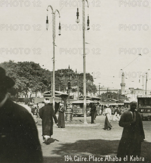 Caire. Place Ahaba el Kabra, View of a square in Cairo, stereograph, number: '194.', inscription: 'These stereo-photographs are being offered for sale and sold wholesale and retail by the ........., ', inscription: 'Submitted.', monogram, address: 'Parklaan 49, HAARLEM, (Holland)', photographer: Richard Sochor, Cairo (city), publisher: De Wereld-Tourist, Haarlem, 1903, Ottoman Empire, North Africa, paper, baryta paper, gelatin silver print, gelatin silver developing print, height 88 mm x width 179 mm, square, circus, A crowded outdoor scene with tall ornate lamp posts and hanging streetlights above a plaza, trees and a minaret visible in the background, a central decorative multi-lantern street lamp, market stalls or small structures with awnings, a tram or streetcar partially visible behind the lamp, numerous people in period clothing and head coverings walking or standing in the foreground, overhead wires crossing the sky.