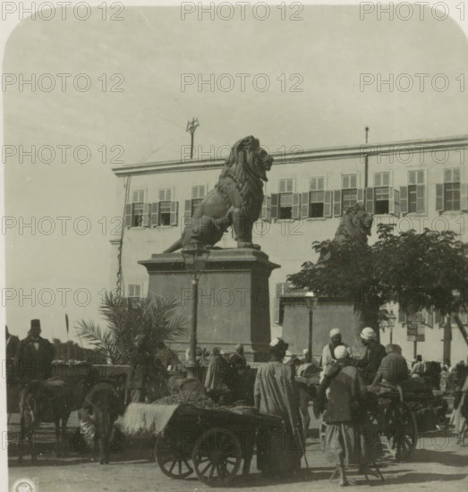 Entrance of Qasr al-Nil Bridge, Cairo, Cairo. Die Brücke von Kasr el Nil, stereograph, number: '6', Qasr al-Nil Bridge, publisher: Neue Photographische Gesellschaft, Steglitz, 1931 - 1935, photographer, Ottoman Empire, North Africa, paper, baryta paper, gelatin silver print, developing gelatin silver print, height 90 mm x width 180 mm, sculpture, bridge, A large stone lion statue sits atop a rectangular pedestal in front of a multi-windowed building, another lion statue is partially visible behind foliage, a lamp post is mounted on the pedestal, people wearing headwraps and cloaks gather and stand around carts and animals, a wooden cart with large spoked wheels and a blanket-draped load is in the foreground, shutters cover many windows of the building, potted palms and trees appear near the base of the pedestal, and the sky fills the upper portion of the image.
