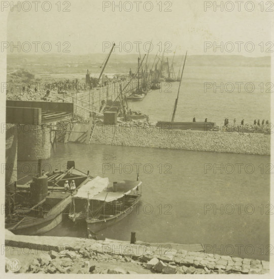 View of the Lower Aswan Dam, Egypt, Nilstaudamm bei Assuan, Upper Egypt, stereograph, number: '39.', number: 'S 20246', Lower Aswan Dam, publisher: Neue Photographische Gesellschaft, Steglitz, 1909 - 1935, photographer, North Africa, Aswan, Lage Aswandam, paper, baryta paper, daylight gelatin silver print, height 88 mm x width 180 mm, barrage, A sepia-toned harbor scene shows a stone quay curving along calm water with multiple small boats moored alongside, several with canvas awnings and visible rigging and masts rising vertically and at angles; a stone wall and piled rocks form the quay edge with a group of people standing and walking along it, and further back more people and construction materials are visible on the shore; cranes and wooden beams extend over the quay and boats, and a larger structure with stone piers projects into the water at the left; distant low hills form the background under a pale sky.