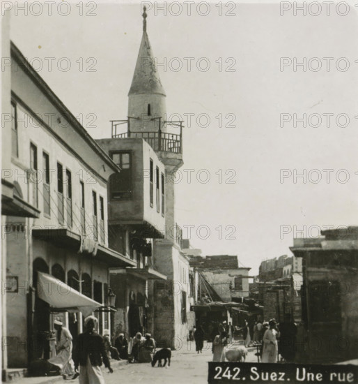Suez. Une rue, View of a street in Suez, stereograph, address: 'Parklaan 49, HAARLEM, (Holland)', inscription: 'Déposée.', monogram, inscription: 'These stereo photographs are marketed and sold wholesale and retail by the ........., ', number: '242.', photographer: Richard Sochor, Suez, publisher: De Wereld-Tourist, Haarlem, 1903, North Africa, paper, baryta paper, gelatin silver print, developing gelatin silver print, height 88 mm x width 179 mm, street, A narrow street scene with multi-story buildings lining the left side, featuring balconies, awnings, and shuttered windows, and a tower with a conical roof and a small balcony near the top rising above the rooftops; people in traditional clothing walk and stand along the street, some clustered near shopfronts, and a few animals are visible on the ground; awnings and street lamps project from the buildings, and the overall image has a monochrome, aged appearance.