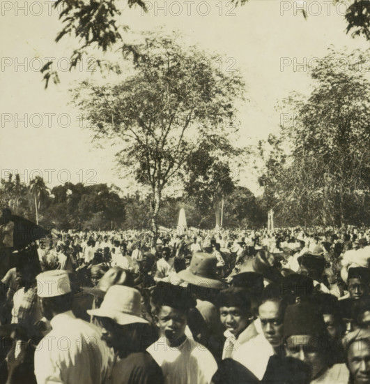 Crowd of people in a square, possibly on Borneo, Indonesia, stereograph, 1900 - 1940, photographer, paper, pasteboard, baryta paper, gelatin silver print, height 90 mm x width 178 mm, crowd, mob, A crowded outdoor scene with many people gathered closely together in the foreground and extending into the middle ground, several wearing hats of different shapes and light-colored clothing; a tall tree with sparse foliage rises behind the crowd, flanked by other trees and dense vegetation, and an obelisk-like monument is visible among the people in the distance; the sky above is light and mostly clear with a few overhanging branches visible at the top edges.