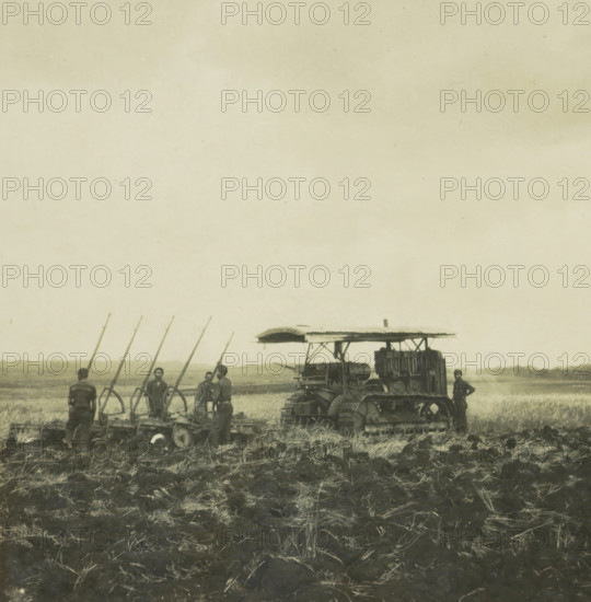 Ploughing machine with farm laborers, possibly on Borneo, Indonesia, stereograph, 1900 - 1940, photographer, paper, pasteboard, baryta paper, gelatin silver print, developed gelatin silver print, height 90 mm x width 179 mm, ploughed field, A wide, sepia-toned outdoor scene showing a field with rough, clumped soil in the foreground and taller grasses beyond, several men standing beside and operating a row of agricultural implements with long vertical poles and wheel-like components, and a large tracked tractor-like machine with a canopy and visible engine components positioned to the right, with one man standing near its rear; the horizon and distant low vegetation are visible under a pale sky.