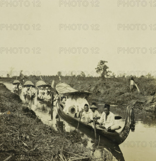 Some boats on the Sungai Martapura river, with both Europeans and original inhabitants of Borneo, stereograph, inscription: 'Terrain exploration windward right on Martapoera river?', Sungai Martapura, 1900 - 1940, photographer, Borneo, paper, pasteboard, baryta paper, gelatin silver print, developing gelatin silver print, height 90 mm x width 179 mm, river, A narrow waterway runs from the foreground into the distance with a line of long wooden boats packed with people sitting and holding paddles, some boats having canopies and patterned fabric draped at the bow, and several passengers wearing hats and light-colored clothing; the banks are lined with grass and reeds, a few small flags are placed along the edge, and a distant tree line is visible beneath a wide pale sky.