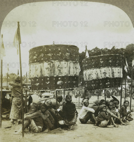 Natives at a festival in Jokjacarta. Java, Javanese people at a festival in Jogjakarta, stereograph, copyright notice: 'Copyright 1902 by CH Graves.', number: '35', number: '95.', photographer: Carleton Harlow Graves, Jogjakarta, photographer: Universal Photo Art Company, Philadelphia (possibly), 1902, North America, Java, Central Java, paper, pasteboard, baryta paper, gelatin silver print, developing gelatin silver print, height 89 mm x width 179 mm, public festivities, A group of people sitting and squatting on the ground under large patterned fabric canopies mounted on poles, with several tall poles bearing plain flags rising above them; many of the seated individuals wear headwraps and patterned clothing and some look toward the camera while others face away; the canopies show layered decorative bands and small motifs, and trees form a distant backdrop beneath a pale, slightly mottled sky.
