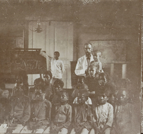 School class with teacher in Manokwari, Indonesia, Dorei. School, Collection D, Geographical gallery of images, collected by MJ Broekhuijsen, headmaster of a school in Amsterdam, stereograph, number: '17.', inscription: '(See pl. 16.) In the background by the blackboard stands the guru, (native teacher, ) who has been appointed by the missionaries in the school also founded by them., ', Manokwari, publisher: Merkelbach & Co., Amsterdam, compiler (person in literature): Martien Johan Broekhuijsen, 1900 - 1902, photographer, compiler (person in literature), paper, pasteboard, baryta paper, daylight gelatin silver print, height 89 mm x width 177 mm, elementary school, class, form, A faded sepia photograph showing a group of children seated in rows in front of a man who stands holding a smaller child, with another child standing beside a chalkboard and a tall door behind them; the room includes a hanging light fixture near the ceiling and a framed map or picture on the wall, and the image ...