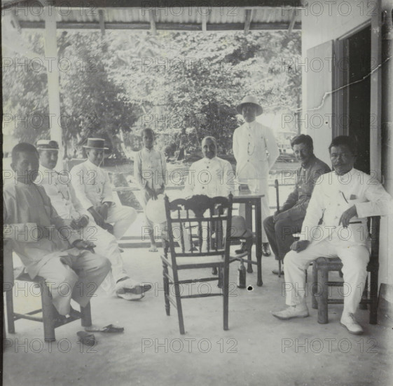 Manoekwari, Seven men and a child on a veranda in Manokwari, West Papua, stereograph, Manokwari, 1900 - 1940, photographer, paper, pasteboard, baryta paper, gelatin silver print, developing gelatin silver print, height 87 mm x width 178 mm, loggia, verandah, porch, A group of men and a boy sit and stand on a covered porch with wooden posts and a corrugated roof, surrounded by leafy trees in the background; several men wear light-colored clothing and some wear hats including a boater and a pith helmet, while others wear darker shirts; wooden chairs and a small table with glassware sit in the center of the scene, one hat rests on a chair and another on the floor, and shoes are visible near the chairs; the porch floor shows scuff marks and a small dark spot, and a doorway or open wall is visible at the right edge of the image.