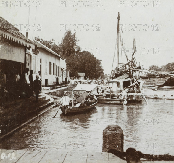 Harbor and toll office in Surabaya, Soerabaja. Small tree, Geographical gallery of images, collected by MJ Broekhuijsen, headmaster of a school in Amsterdam, Collection D, stereograph, number: 'D.19.', inscription: 'De Boom is the landing place for small vessels (such as praus) coming from the large boats to the toll office, ', Surabaya, publisher: Merkelbach & Co., Amsterdam, compiler (person in literature): Martien Johan Broekhuijsen, 1900 - 1902, photographer, compiler (person in literature), Java, East Java, paper, pasteboard, baryta paper, daylight collodion silver print, height 88 mm x width 177 mm, harbour, toll gate, A riverside scene showing a row of low buildings with tiled roofs and awnings along a stepped stone quay, several people standing on the steps and by the building, a small wooden boat with a person rowing in the water near the quay, larger boats with masts and rigging moored along the opposite side, tarpaulins and canopies covering parts of the boats, trees and fol...