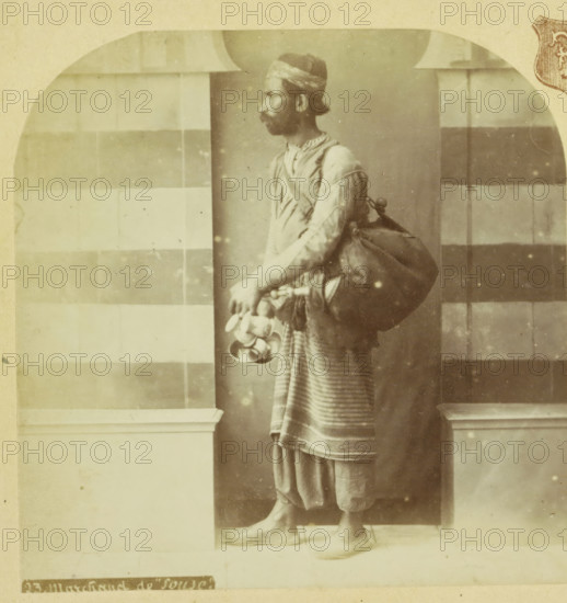 Koopman te Sousse, Tunisia, Marchand de 'Souse', Palestine et Syrie, Palestine and Syria, stereograph, number: '23.', photographer: F & E, Sousse, Paris (possibly), 1879 - 1910, North Africa, pasteboard, paper, albumen print, height 87 mm x width 173 mm, merchant, salesman, A sepia-toned photograph of a man standing in profile, wearing a headwrap and a long sleeved tunic with a striped lower garment, a large bag slung over his shoulder, and holding a cluster of round objects and shallow bowls in one hand, positioned between two architectural pillars with a checkered wall pattern behind him and wearing pointed shoes while standing on a tiled floor.
