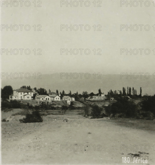 View of Jericho, Jérico, stereograph, address: 'Parklaan 49, HAARLEM, (Holland)', inscription: 'These stereo-photos are being brought into trade and sold wholesale and retail by the ........., ', monogram, inscription: 'Submitted.', number: '180.', photographer: Richard Sochor, Jericho, publisher: De Wereld-Tourist, Haarlem, 1903, Palestine, West Bank, paper, baryta paper, gelatin silver print, developed gelatin silver print, height 88 mm x width 179 mm, city-view in general, 'veduta', A monochrome photograph showing a wide sandy or gravelly foreground leading to a slightly elevated area with scattered low shrubs and vegetation, behind which a row of small buildings with pitched roofs and several larger multi-story houses sit among tall, narrow trees and other foliage, with a distant low ridge or hill visible across the middle of the image and a pale, textured sky occupying the upper portion.