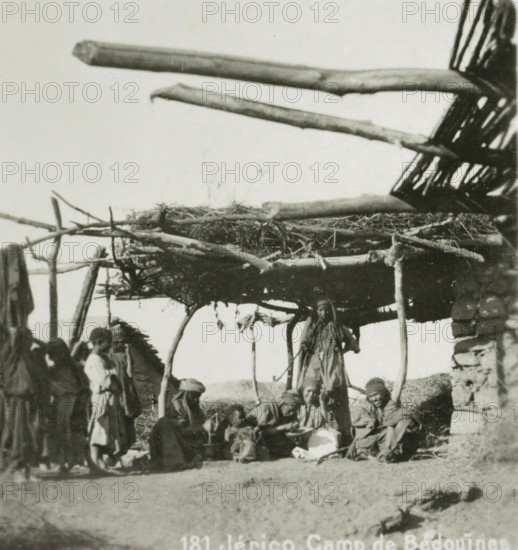 Bedouin camp in Jericho, Jérico. Camp of the Bedouins, stereograph, number: '181.', address: 'Parklaan 49, HAARLEM, (Holland)', monogram, inscription: 'These stereo photographs are being brought into trade and sold wholesale and retail by the ........., ', inscription: 'Submitted.', photographer: Richard Sochor, Jericho, publisher: De Wereld-Tourist, Haarlem, 1903, Palestine, West Bank, paper, baryta paper, gelatin silver print, developing gelatin silver print, height 86 mm x width 180 mm, tent-camp, wandering communities: nomads, A group of people wearing layered clothing and head coverings are gathered beneath a simple shelter made of rough wooden poles and a woven roof, with several large horizontal logs projecting outward above; some individuals are standing at the shelter's edge while others sit or crouch on the ground and on low stone structures, and portions of a stacked stone wall are visible at one side.