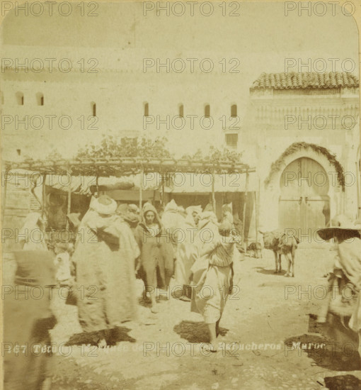 Tetuan. Mosque de la place des Babucheros. Maroc, Passers-by on the Babucheros Square in Tétouan, stereograph, number: '264', number: '167', Tétouan, publisher: American Stereoscoop Platen Gezelschap Kunst Vermaak en Onderzoek, Netherlands, 1879 - 1910, American Stereoscoop Platen Gezelschap Kunst Vermaak en Organisatie, photographer, North Africa, pasteboard, paper, albumen print, albumendruk, height 88 mm x width 178 mm, square, circus, A faded street scene showing a group of people clustered in front of a light-colored wall with a row of small arched openings and a tiled roof section at one end, some figures wearing long robes and wrapped head coverings and a few faces visible among them, a wooden canopy or stall structure with branches and foliage overhead behind the crowd, an arched doorway set into the wall decorated with a garland and secured by a horizontal bar, a load-bearing animal standing near the doorway with packs on its back, and a ground surface showing shadows and unev...