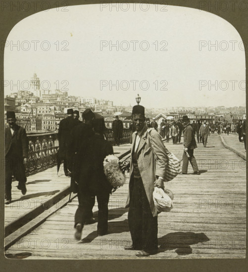 Galata, the Foreign part of Constantinople, from the Outer Bridge, Fan seller on the Galata Bridge in Istanbul, stereograph, copyright notice: 'Copyright 1901 by HC White Co.', number: '3', number: '4309.', inscription: 'The ' PERFEC' STEREOGRAPH. (Trade Mark.), Patented April 14, 1903. Other Patents Pending.', Galatabrug, publisher: HC White Co., New York (city) (possibly), 1901, photographer, Istanbul, North America, paper, pasteboard, baryta paper, gelatin silver print, developing gelatin silver print, height 88 mm x width 177 mm, pontoon bridge, Galata Bridge, A sepia-toned photograph of a wooden pedestrian bridge crowded with people, showing a foreground man facing the camera wearing a long coat and a hat, holding stacks of round objects and a patterned umbrella, with another man walking away beside him wearing dark clothing and a fez, ornate metal railings along the left side of the bridge, a row of people walking in both directions further along the bridge, a distant cityscape o...