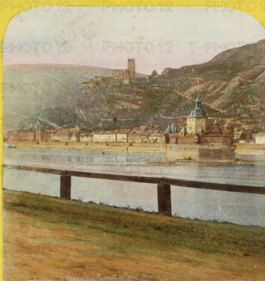 View of Kaub and the Burgen Gutenfels en Pfalzgravenstein, Germany, Pfalz, Caub et ruines du Gutenfels, Bords du Rhin, Allemagne, stereograph, inscription: 'Déposé.', number: 'N. 470', photographer: Hippolyte Jouvin, Kaub, 1860 - 1870, Rhineland-Palatinate, pasteboard, paper, albumen print, height 86 mm x width 177 mm, landscape with ruins, city-view, and landscape with man-made constructions, Burg Gutenfels, Burg PfalzGrafstein, A landscape scene showing a river in the middle ground with a long bridge crossing it supported by dark vertical piers, a grassy foreground sloping down toward the water, and a distant town along the far bank featuring rows of buildings and a prominent tower with a domed roof and spires; beyond the town are terraced hills rising into the background with a ruinous tower on a hilltop, all beneath a pale, lightly mottled sky.