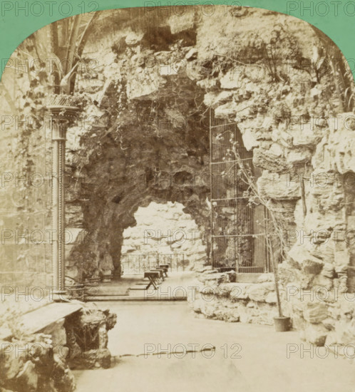 Freshwater section of the Berliner Aquarium, seen from the aviary, Die Süsswassergalerie von der Voliere aus., Galerie d'eau douce vue de la volière, Berliner Aquarium, Aquarium de Berlin, stereograph, number: '16.', photographer: SP Christmann, Berlin, publisher: SP Christmann, Berlin (possibly), 1869 - 1890, SP Christmann, photographer, publisher, pasteboard, paper, albumen print, height 86 mm x width 176 mm, zoological garden, zoo, seen as place where non-domestic animals are kept, Unter den Linden, A sepia-toned scene showing a rocky archway formed by rough stone walls with a narrow paved pathway beneath, a metal column with decorative ridges and a lattice planter attached standing to the left, a metal gate with vertical bars set into the stone on the right, small wooden benches arranged along the path under the arch, irregular stones and rock outcroppings lining both sides, thin climbing plants and vines trailing over the stones, and a tall slender potted plant with a thin trunk p...
