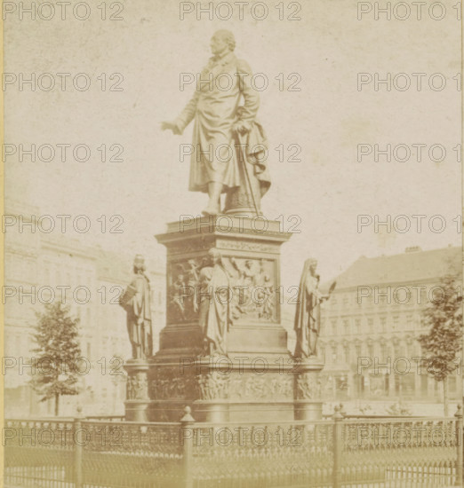 Das Stein-Denkmal, Statue of Baron van Stein in Berlin, Berlin and Environment, stereograph, photographer: Sophus Williams, Berlin, after sculpture by Hermann Schievelbein, 1877, Hermann Schievelbein, after sculpture by, pasteboard, paper, albumen print, height 86 mm x width 176 mm, Person, Heinrich Friedrich Karl vom und zum Stein, monument, statue, Dönhoffplatz, A tall stone statue of a robed man stands on a multi-tiered ornate pedestal surrounded by additional robed figures and sculpted relief panels, with a wrought iron fence in the foreground and large classical buildings and trees visible in the background.