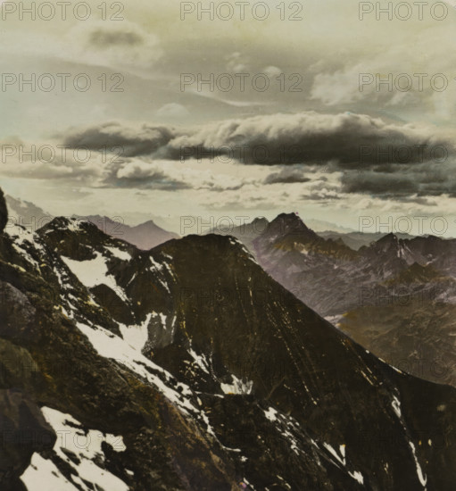Allgäu Alps. Sonnenuntergang am Bockar-Kopf (2609 m), Bockar-Kopf in the Allgäu Alps, Germany, Deutschland. Bayern, stereograph, monogram: 'BW', inscription: 'Deutschland. Bayern. Allgäu Alps. Sonnenuntergan, am Bockar-Kopf (2609 m)., Germany. Bavaria. Allgäu-Alps. Sunset at the 'Bockarkopf' (2609 m)., Allemagne. Vacière. Les Alpes de l'Allgäu. Coucher du, soleil au 'Bockarkopf' (2609 m)., Alemania. Baviera. Los Alpes del, Allgäu. Puesta del sol al 'Bockarkopf' (2609 m).', number: '53', photographer: BW, Allgäuer Alpen, 1898 - 1935, BW, photographer, Alpen, paper, gelatin silver print, hand coloring, developing gelatin silver print, height 88 mm x width 169 mm, mountain-range, snow peaks, Alps, Allgäu Alps, A rugged mountain landscape fills the image with dark rocky peaks and ridges in the foreground showing patches of snow clinging to steep slopes, while more distant jagged mountains fade into softer tones; above them a wide sky is dominated by layered clouds, including a large, elong...